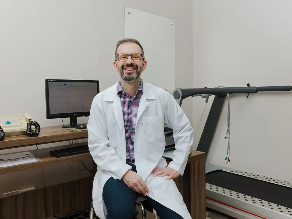 Dr. Marcos Sardinha na sala de exames, ao lado do aparelho de eletrocardiograma e da esteira usada no teste ergométrico